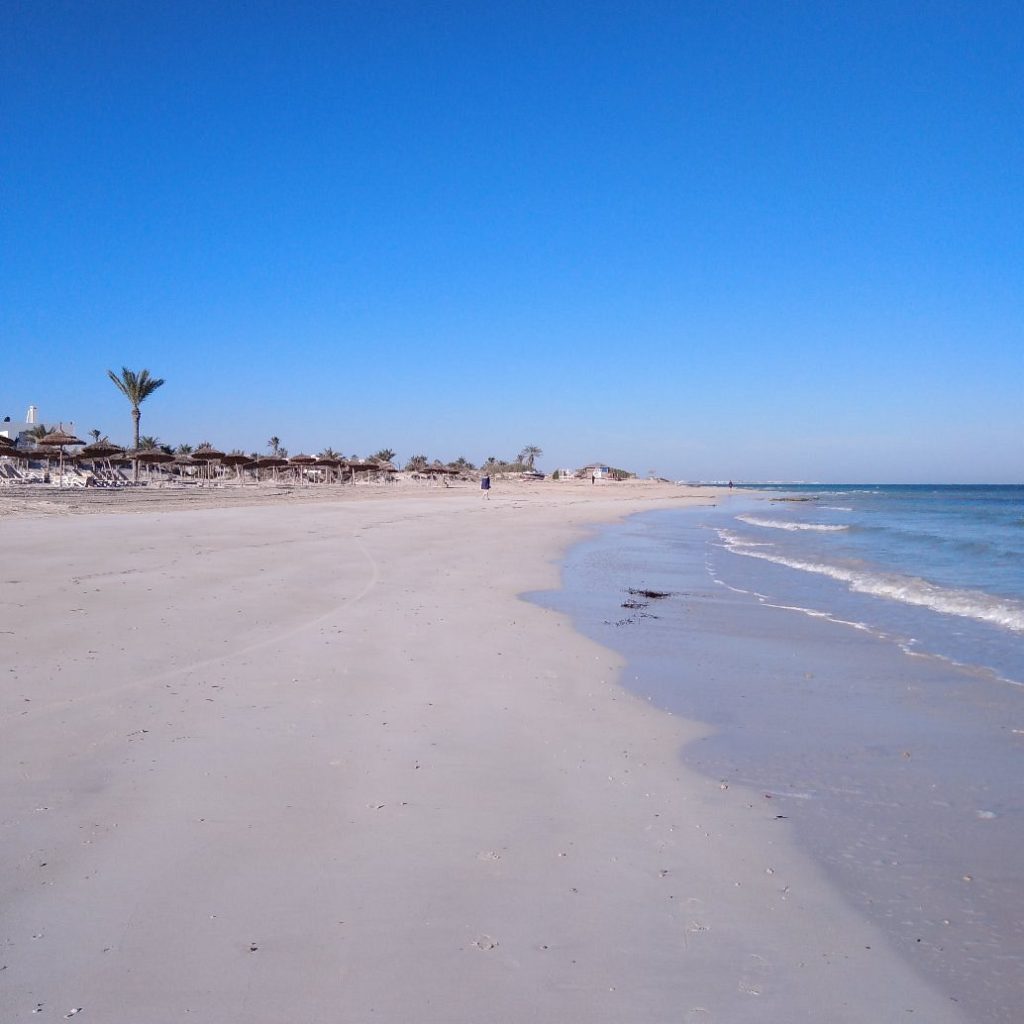 Extensión de arena con mar a la derecha y sombrillas de paja a la izquierda